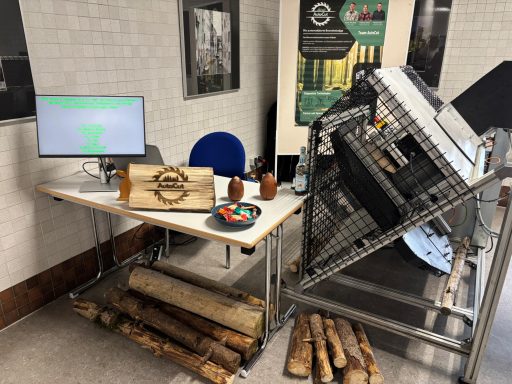 Ein Tisch mit Computer und Werkzeugen, umgeben von Holzstücken und einer großen Maschine.