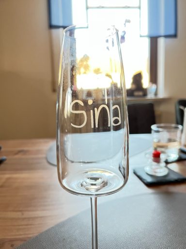 Sektglas mit eingraviertem Namen "Sin" auf einem Tisch.